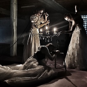 Photo of three people dressed in white wedding gown like dresses, photo has a dark tone filter. Woman lies on floor propped up by a stool and two other figures lean over person one has a candelabra (with three shining lights) the other another item. Photo feels a bit spooky and it looks like people have white painted faces and dark eyemakeup